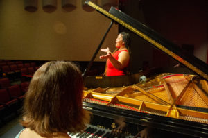 Back wide shot of student playing the piano while another other students perform on stage. HELP