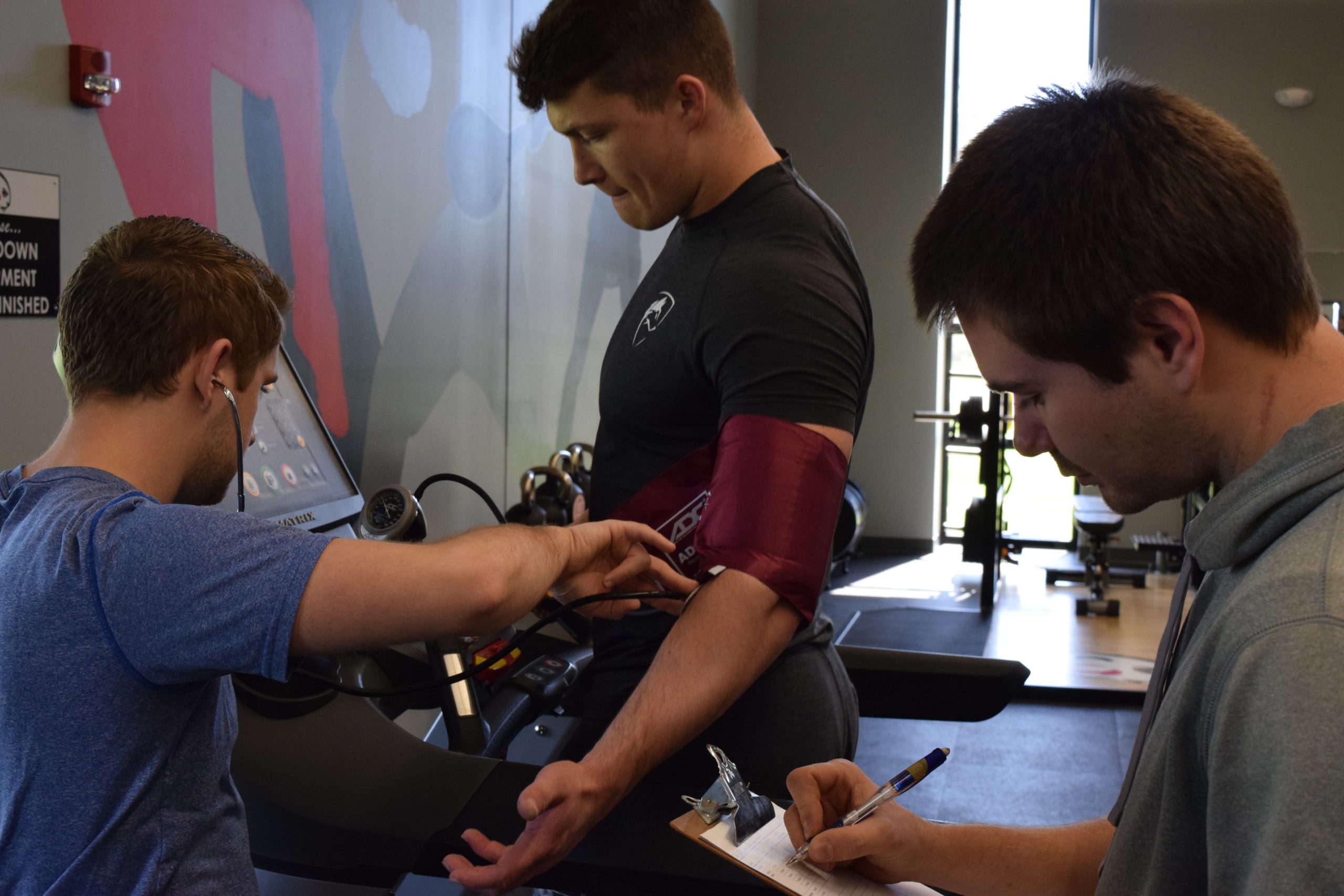 Two Blackburn students analyzing a student-athlete. One of the students uses a sphygmomanometer and a stethoscope on the athlete while the other one takes notes.