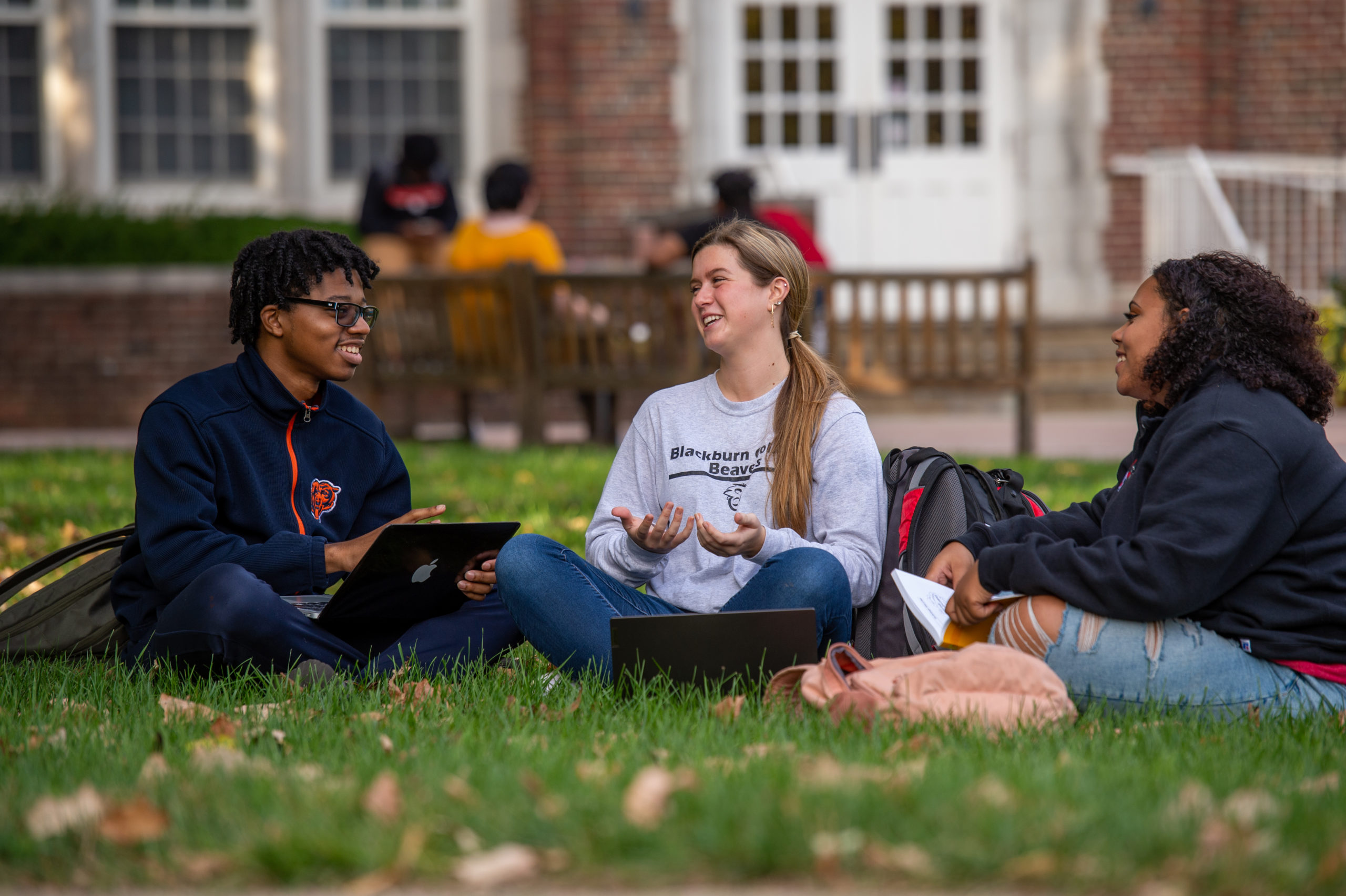 Three Blackburn students sitting and conversating on the grass near Hudson Hall's Quad. A group of students can be seen sitting at a bench in the background.