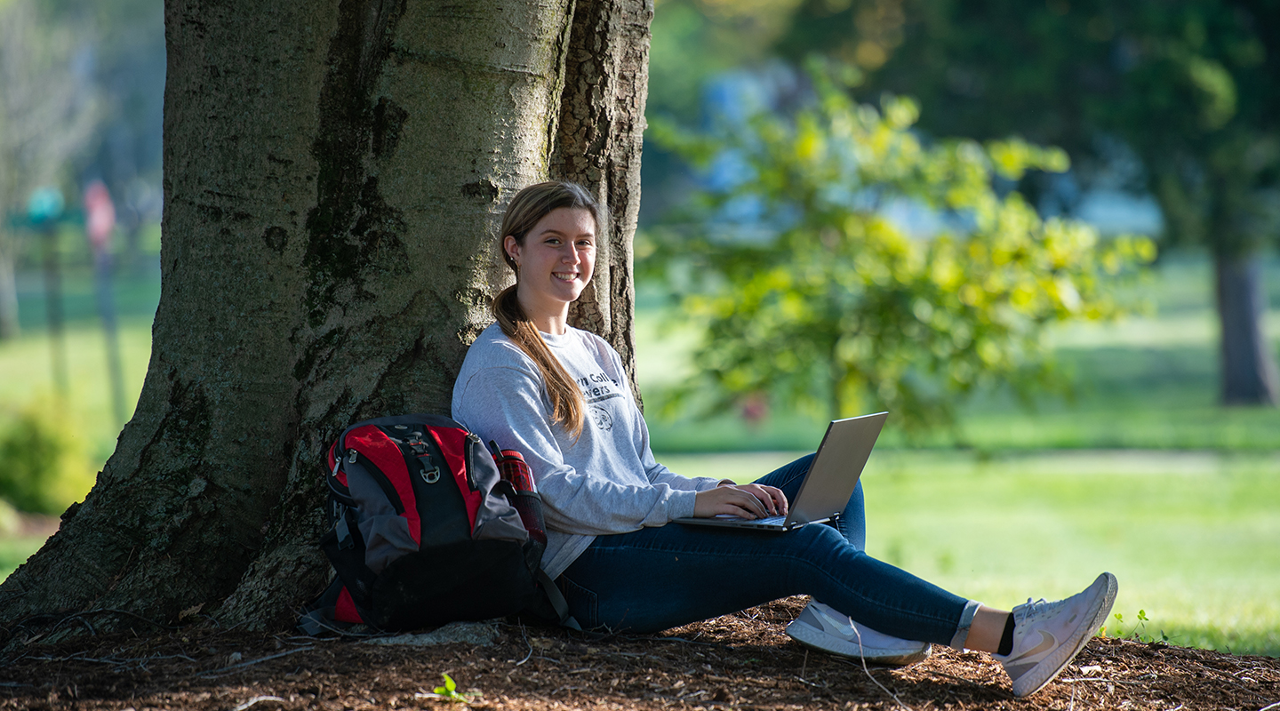 Blackburn student sitting on the ground and leaning against a tree while working on her laptop.
