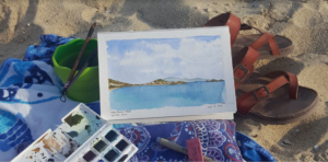Close-up of painting lying on top of a towel, a pair of sandals, and some painting supplies. All of these items are sitting on top of sand.