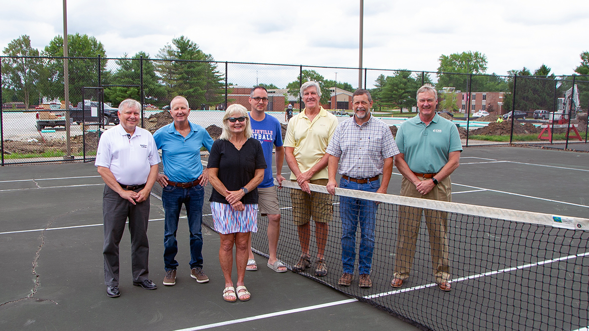 Seven individuals representing Blackburn College and community organization Winning Communities gather on the Blackburn College tennis courts and the future site of the 