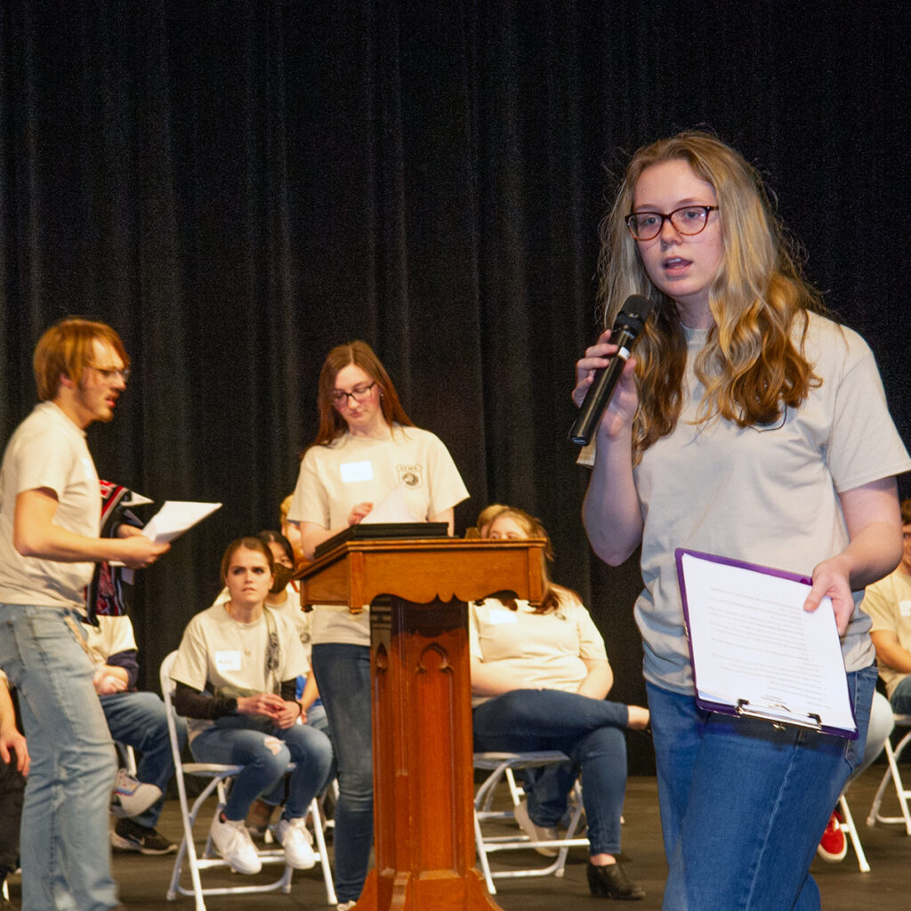 Blackburn College student with microphone and clipboard addresses crowd of high school students, while two other Blackburn students prepare awards behind a podium in the background.,