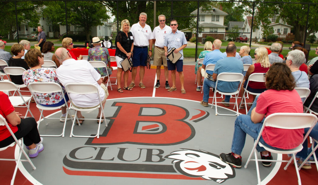 Representatives of the Blackburn Athletic Boosters (B-Club) pose in front of half court on the outdoor basketball court featuring the B-Club logo for sponsoring the court