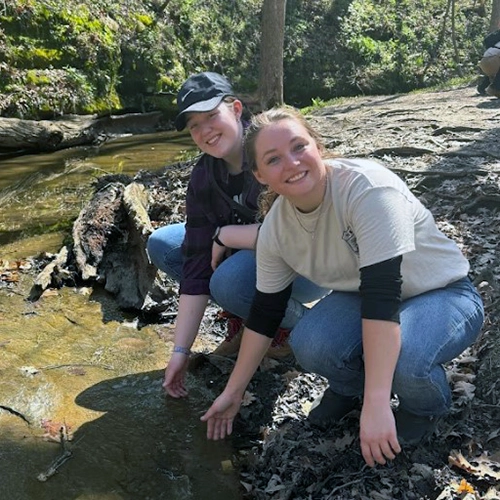 Two women kneel at creek on field study trip