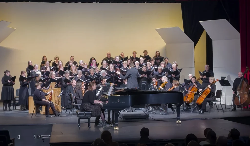 Blackburn's three choirs stand on stage, joined by stringed instruments and a student center stage at a piano performing an original composition