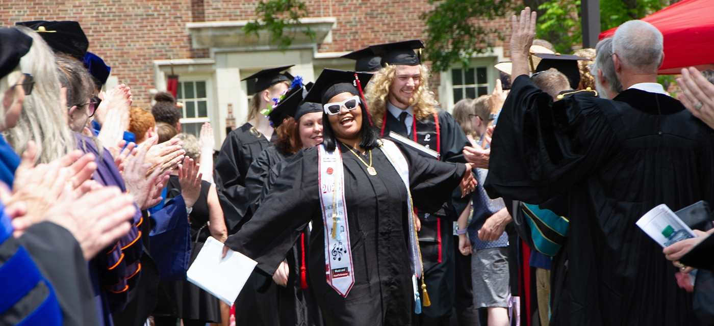 A line of graduates walk down brick pathway surrounded by faculty and staff on each side